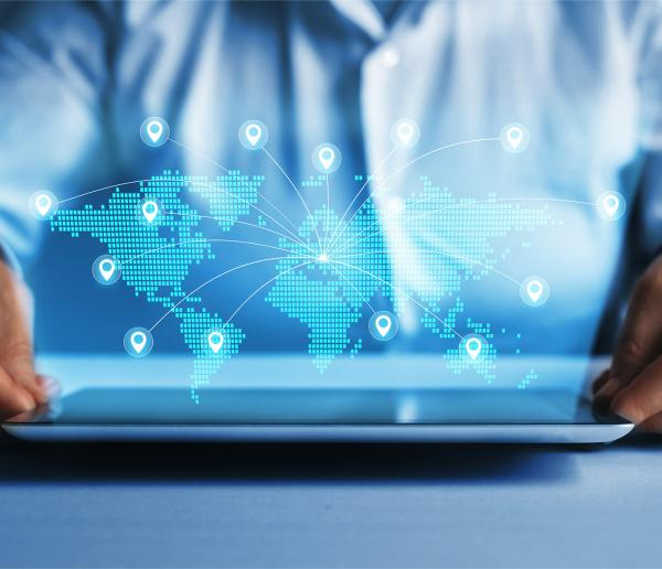 decorative. Person in a white shirt holding a tablet on a table. Above the tablet, a glowing digital world map is displayed with interconnected lines and multiple location markers, symbolizing global connectivity and cybersecurity. The background is dark blue with a futuristic, tech-inspired design
